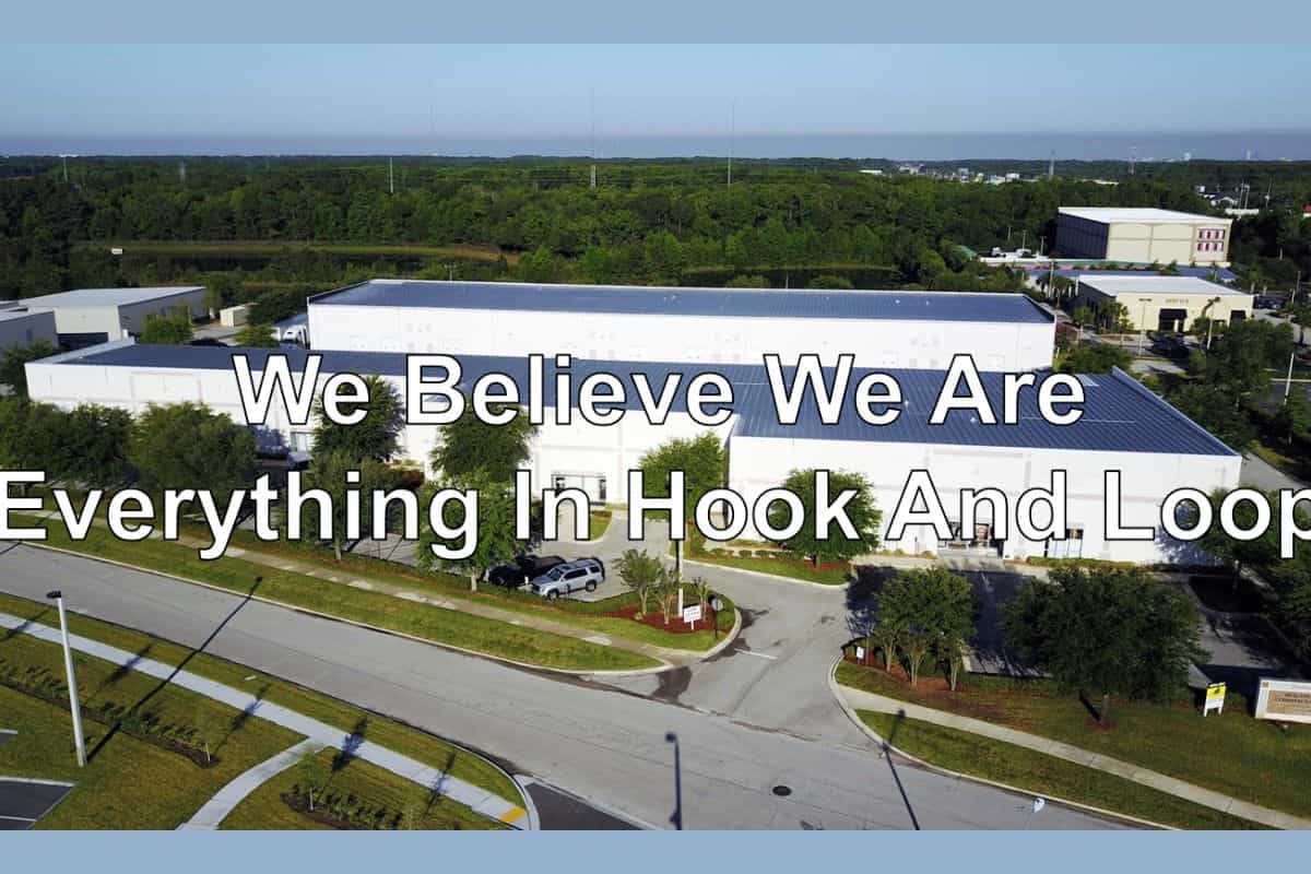 An aerial view of a white commercial warehouse facility surrounded by green trees and asphalt parking lots with text overlay reading We Believe We Are Everything In Hook And Loop for manufacturing Hook and Loop Straps.