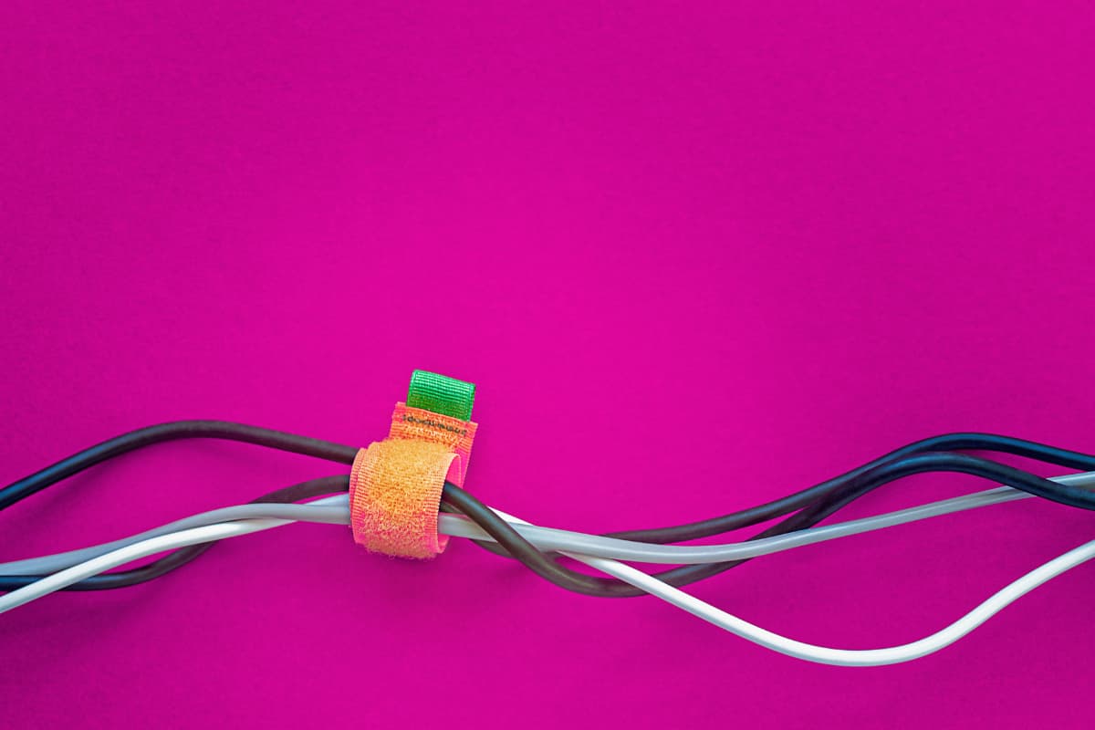 A bundle of electrical cords organized and bundled together on a bright pink background using a colorful green and orange fastener made of Hook and Loop Straps.